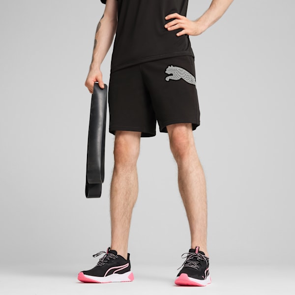 Against grey, a model poses in black shorts, t-shirt, and trainers, holding a band.
